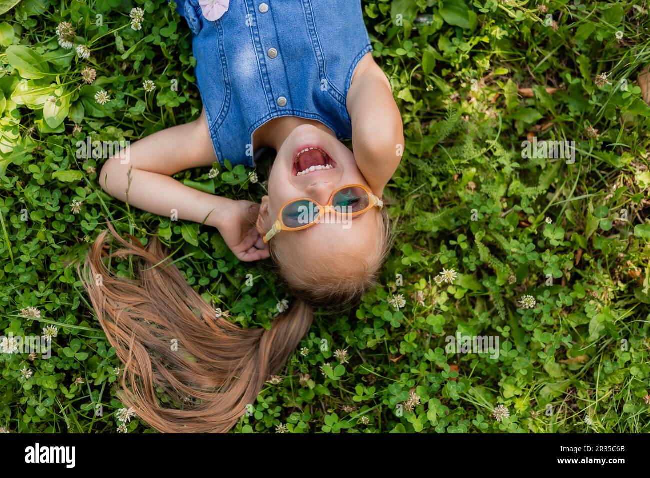 happy little girl lying on the green grass in summer top view. A child with down syndrome wearing glasses for vision. High quality photo Stock Photo