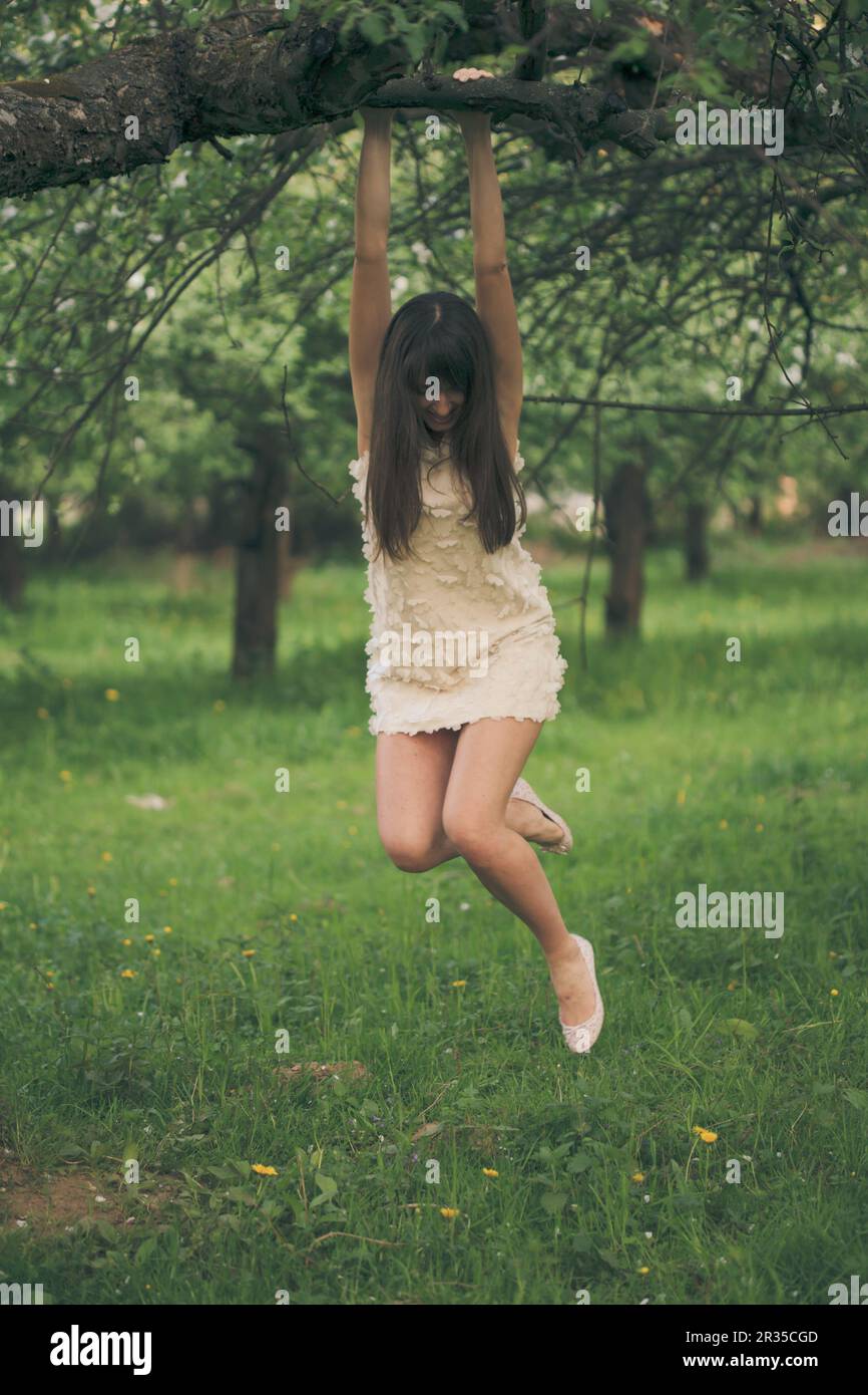 Pretty girl playfully hangs on a tree branch with bent legs Stock Photo