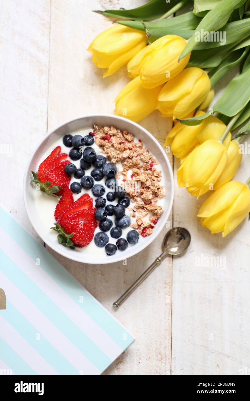 Muesli with yoghurt, blueberries and strawberries next to a bunch of yellow tulips Stock Photo