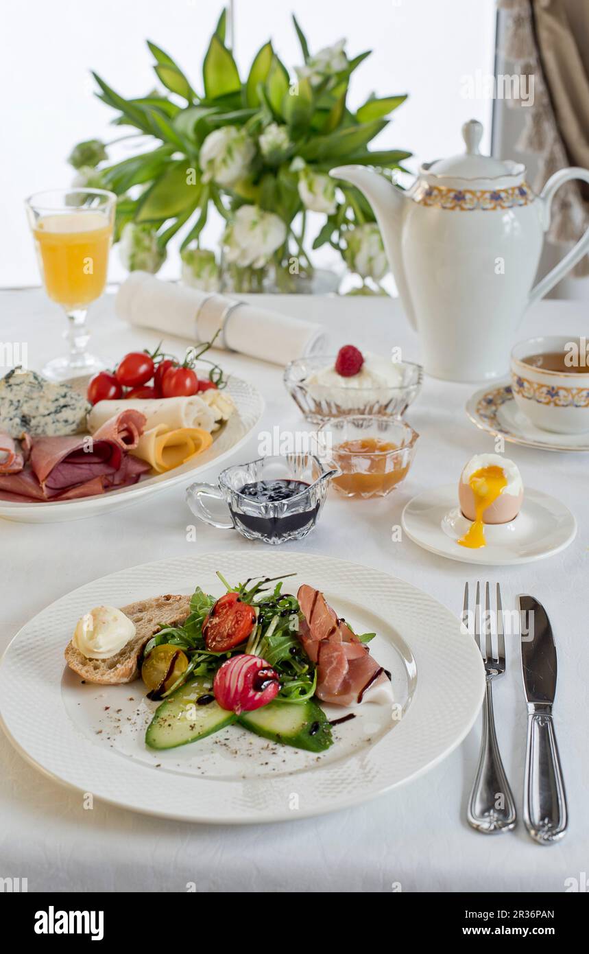 A breakfast table with prosciutto, mixed leaves, balsamic vinegar, bread and butter, cucumber, radishes, a soft boiled egg and a cheese plate Stock Photo