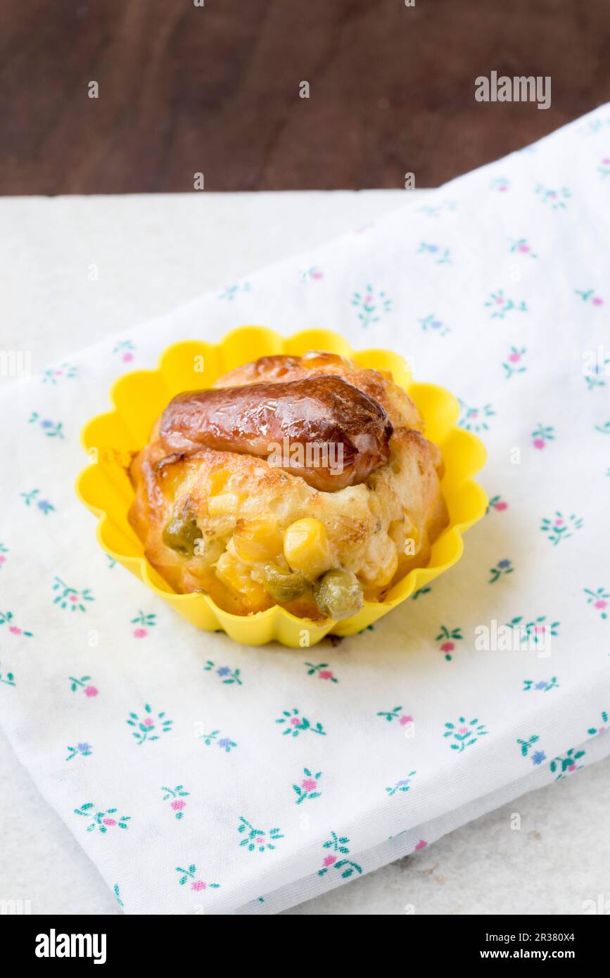 A savoury muffin with a Frankfurter sausage, corn and peas in a yellow muffin case on top of a floral napkin Stock Photo
