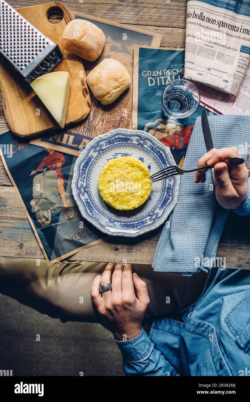 Risotto Milanese on a rustic wooden table with a newspaper, cheese and bread rolls Stock Photo