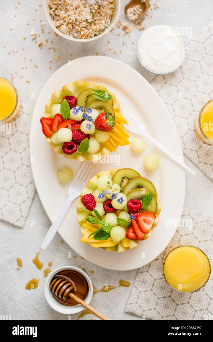 A healthy breakfast: fruit salad served in melon halves, yoghurt, granola, orange juice and honey Stock Photo