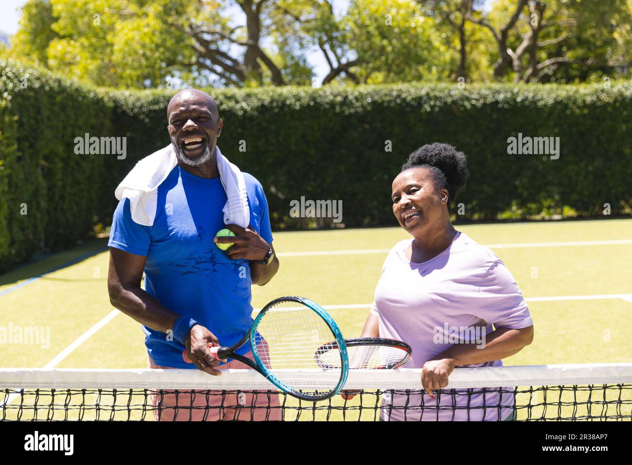 Happy senior african american couple with tennis rackets laughing on sunny grass court Stock Photo