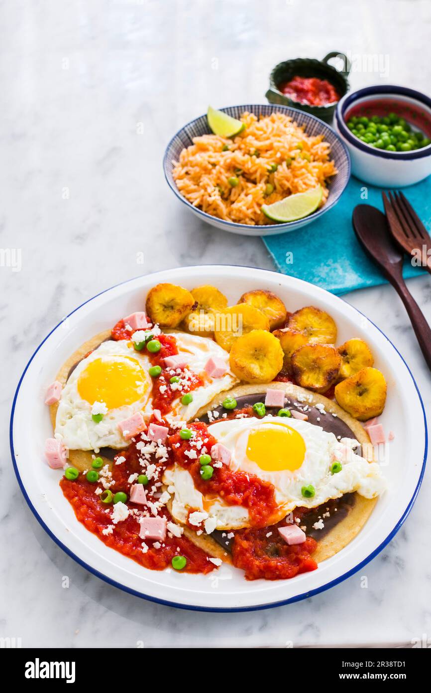 Huevos Motuleños (fried eggs on a tortilla with black bean puree, chilli sauce, plantains, bacon and cheese, Mexico) Stock Photo