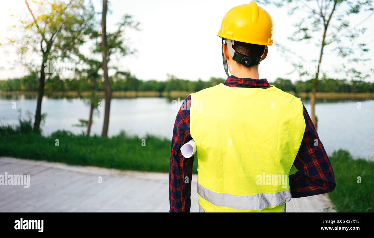 Construction worker wearing yellow vest staying in front of the lake. Worker holding construction plan. Stock Photo