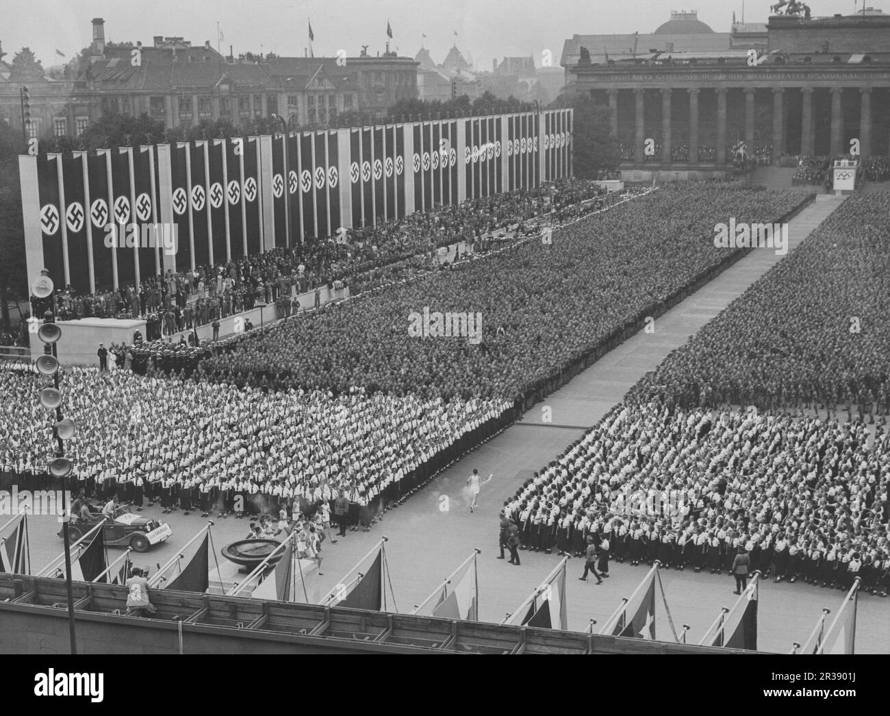Berlin Olympics 1936. The 1936 Summer Olympics (German: Olympische Sommerspiele 1936), officially known as the Games of the XI Olympiad (German: Spiele der XI. Olympiade) and commonly known as Berlin 1936, were an international multi-sport event held from 1 to 16 August 1936 in Berlin, Germany.  Pictured the olympic flame being transported to the Olympiastadion in Berlin by runners, this ceremony is called Olympic torch relay. The nazi flags are seen hanging there. Stock Photo