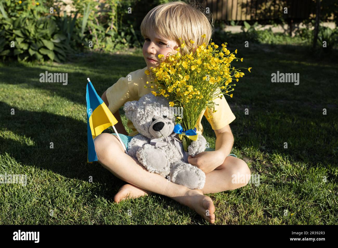 unhappy seven-year-old boy sits on grass with teddy bear, Ukrainian flag, bouquet of yellow flowers. Children against war.concept of peace for Ukraini Stock Photo