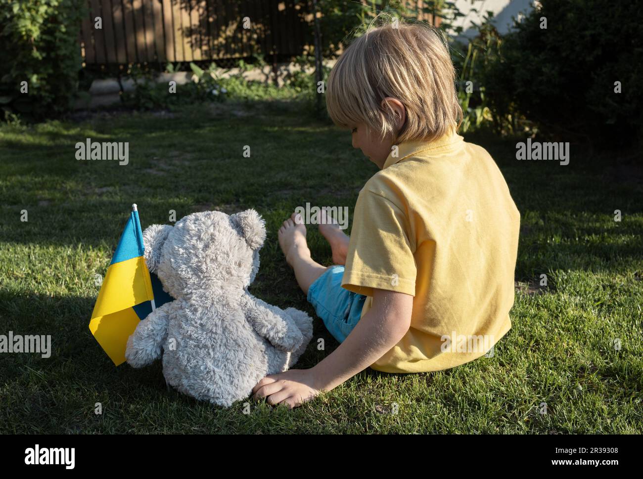 boy and his imaginary friend, a toy bear with a Ukrainian flag, are sitting on the grass with their backs. Children want peace for the Ukrainian peopl Stock Photo