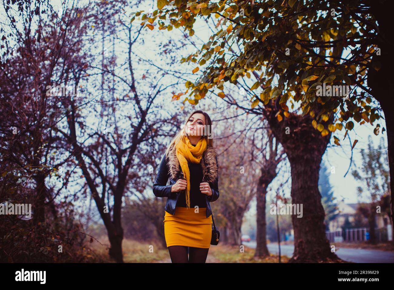 Portrait of young woman wearing black leather jacket and yellow scarf outdoors. Pretty girl walking in autumn park. Stock Photo