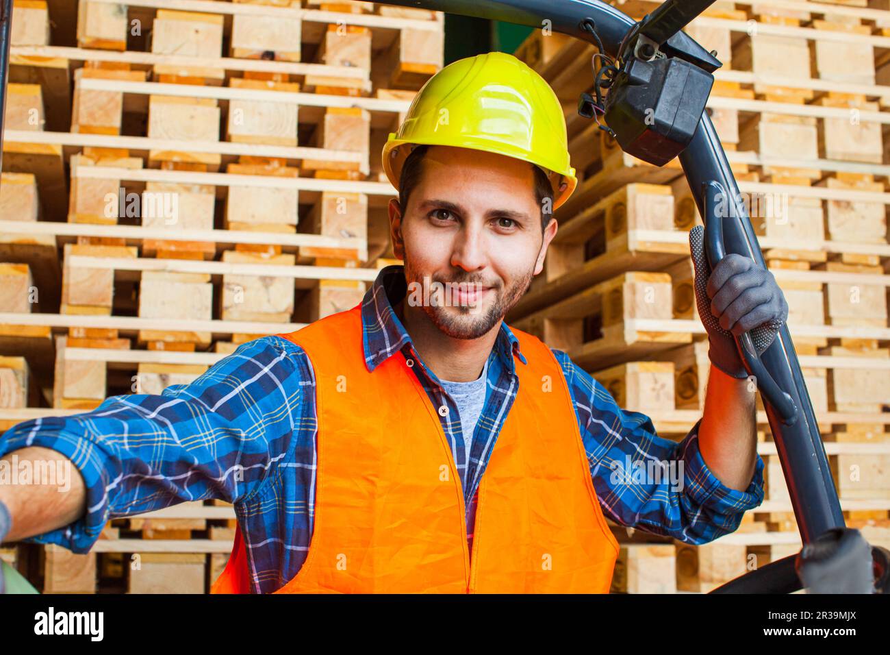 Workman wears protective helmet, vest and gloves. Worker standing near forklift in warehouse. Stock Photo