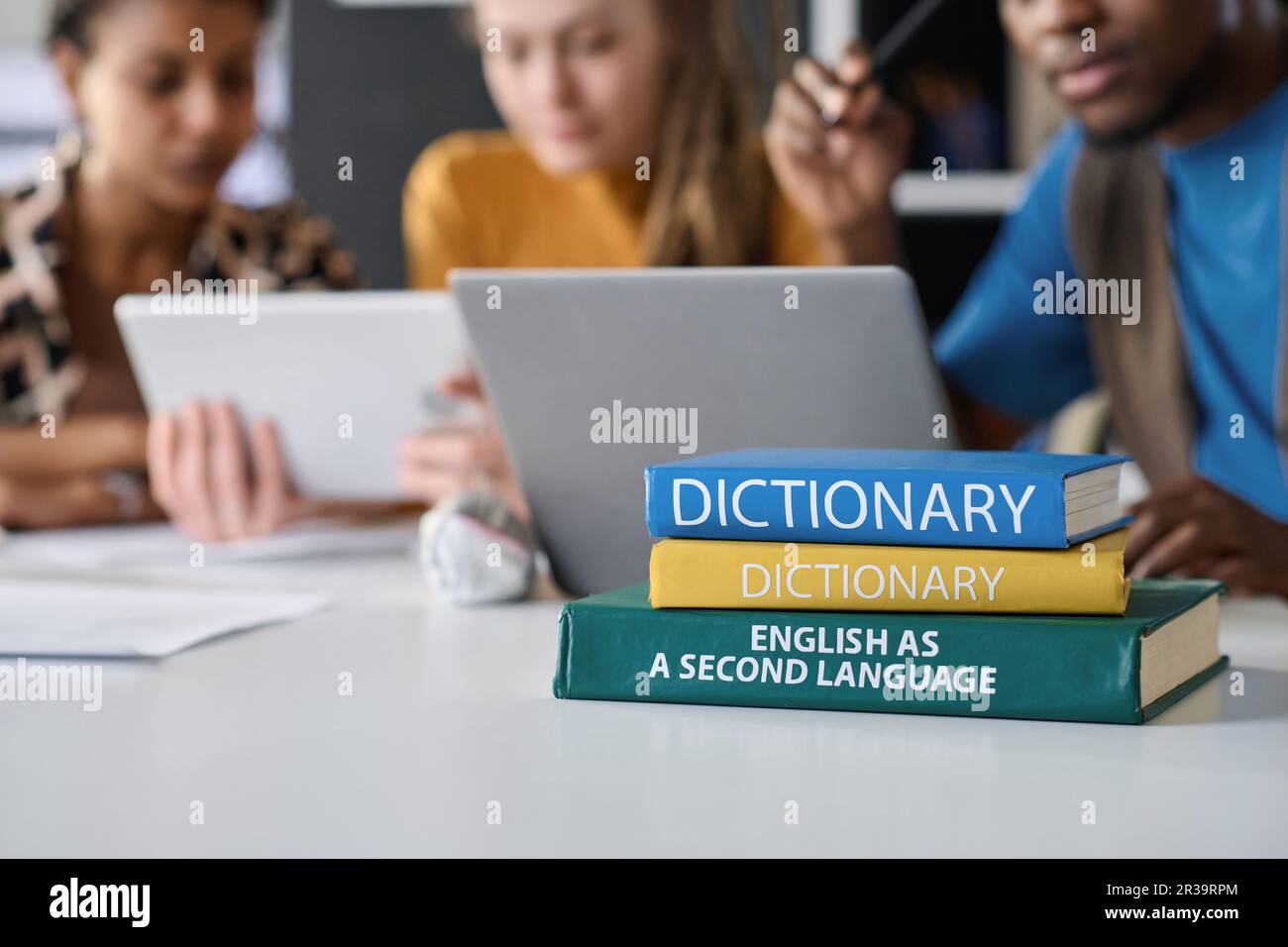 Close-up of stacks of books lying on table with students studying foreign language in background Stock Photo
