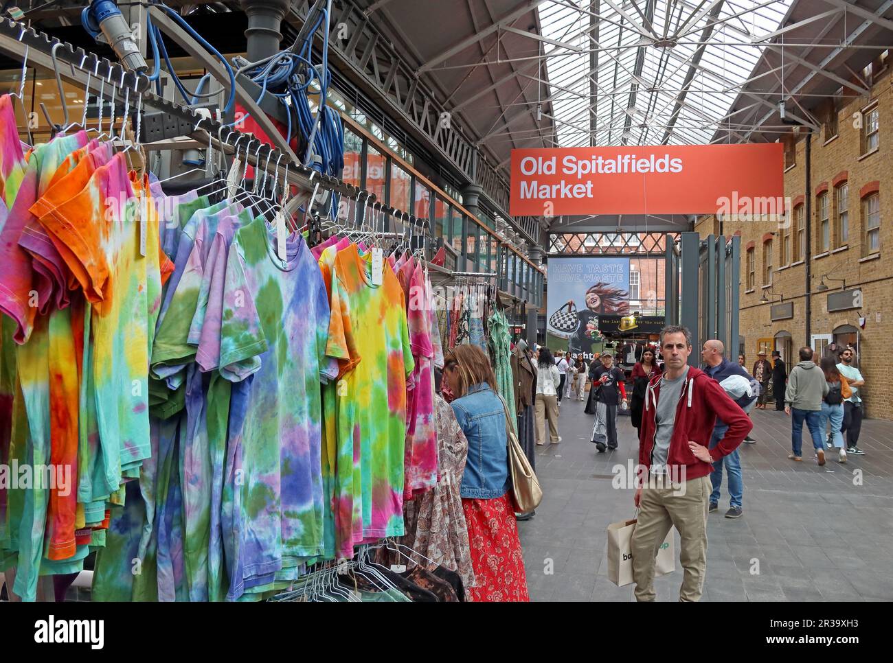 Clothing stalls selling colourful new garments inside the covered hall of Old Spitalfields Market in East London, with shoppers moving inside Stock Photo