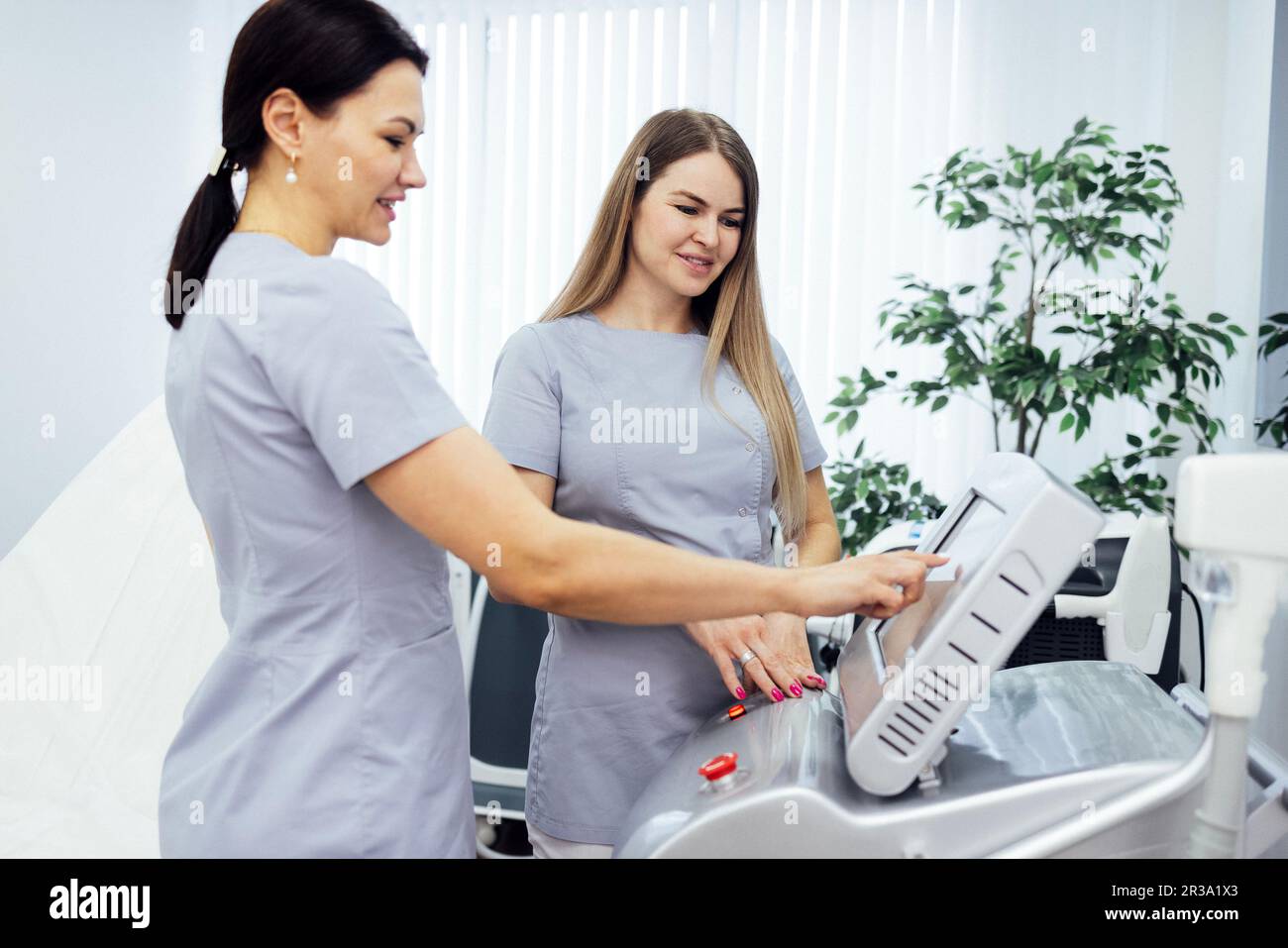 Woman cosmetologist touches screen of cosmetology equipment to set necessary parameters and teaches her colleague about it on beauty salon. Two doctor Stock Photo