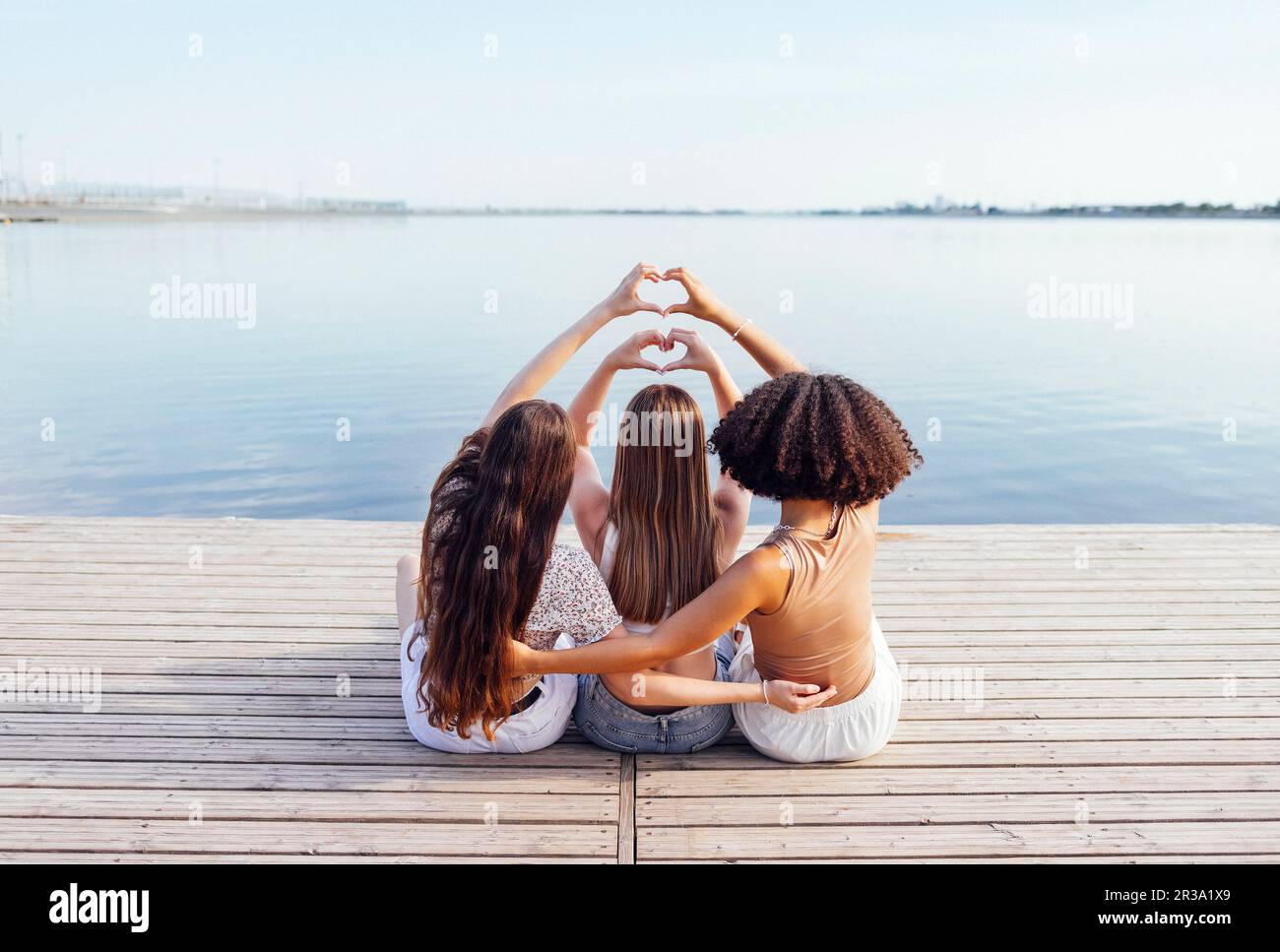 Three female teenagers of different ethnic groups show heart symbols with their hands. Teen girls in casual clothes sit on wooden bridge near the rive Stock Photo