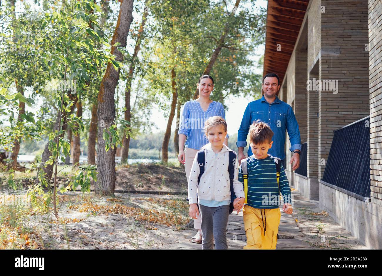 Young parents seeing their cute kids off to school. Smiling mom, dad, daughter and son go down