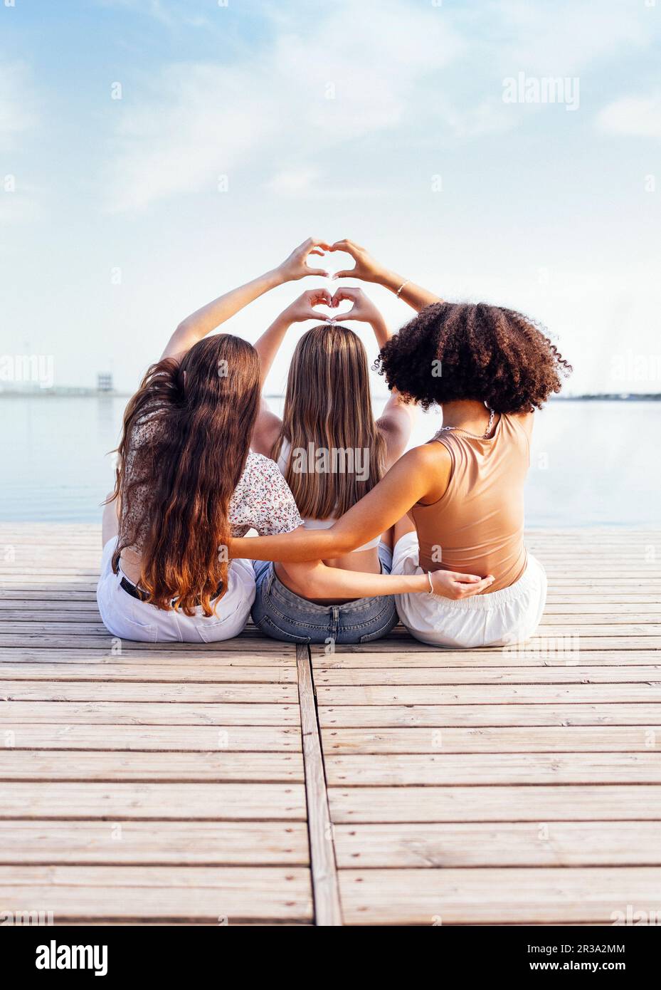 Three female teenagers of different ethnic groups show heart symbols with their hands. Teen girls in casual clothes sit on wooden bridge near the rive Stock Photo