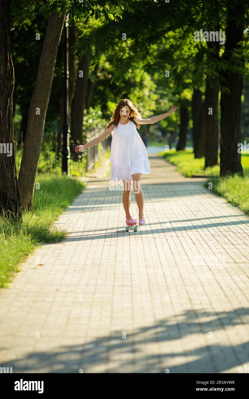 Lovely urban woman in white dress with a pink skate. Young girl riding in the park on a skateboard Stock Photo