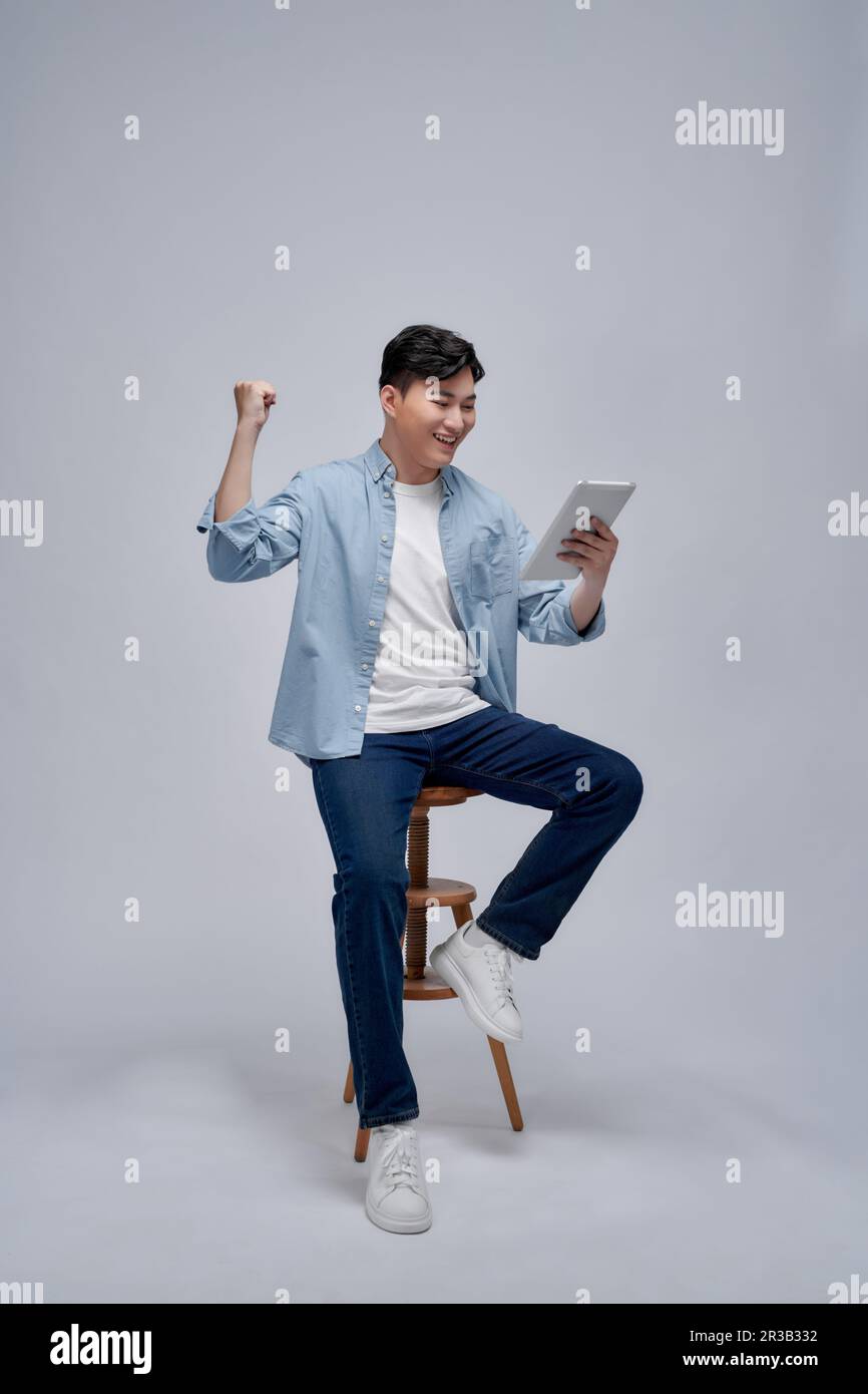 Cheerful casual man holding his tablet and celebrating with his fist in the air while sitting on a chair Stock Photo