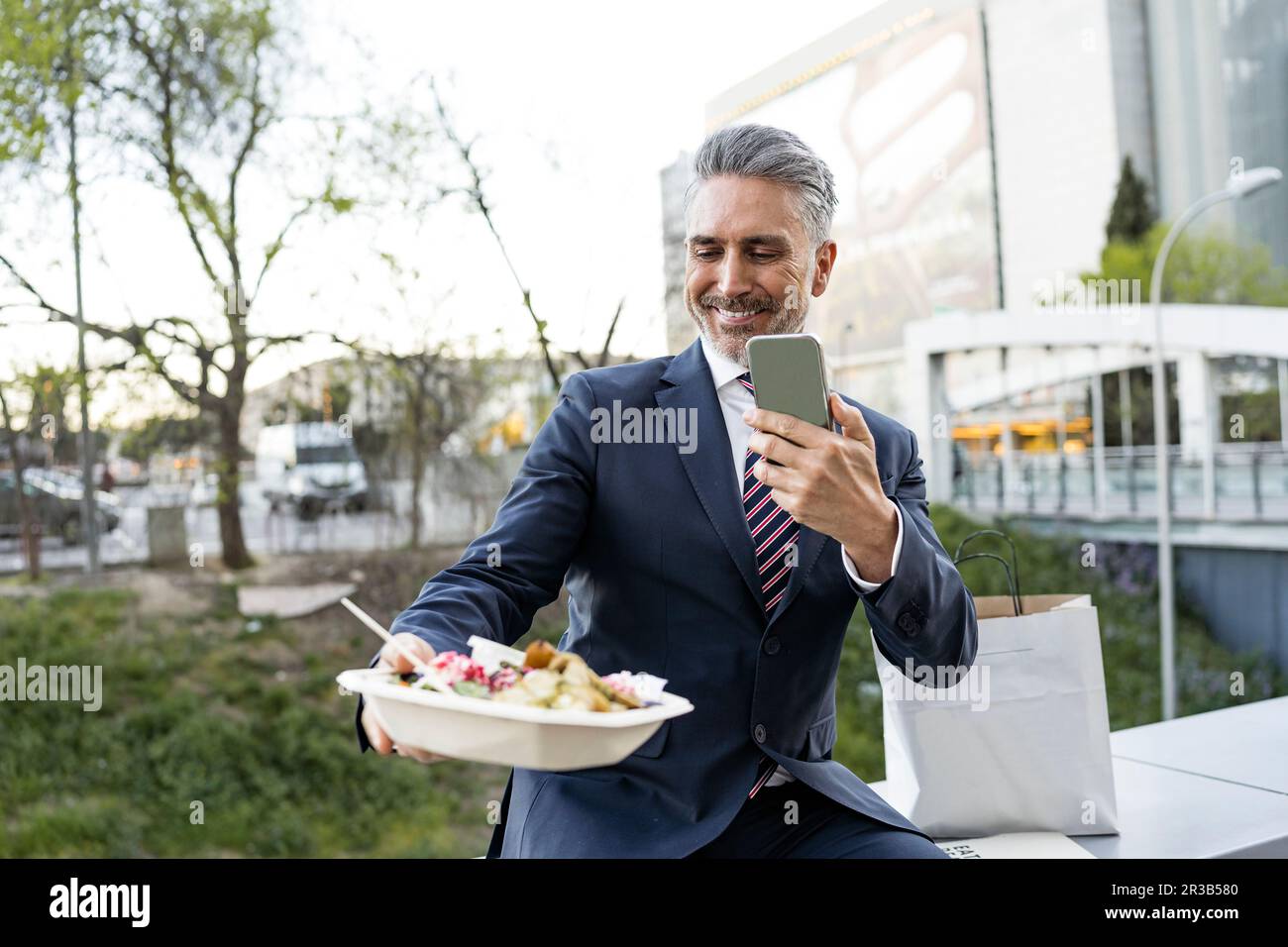 Businessman photographing meal box through smart phone sitting on wall Stock Photo