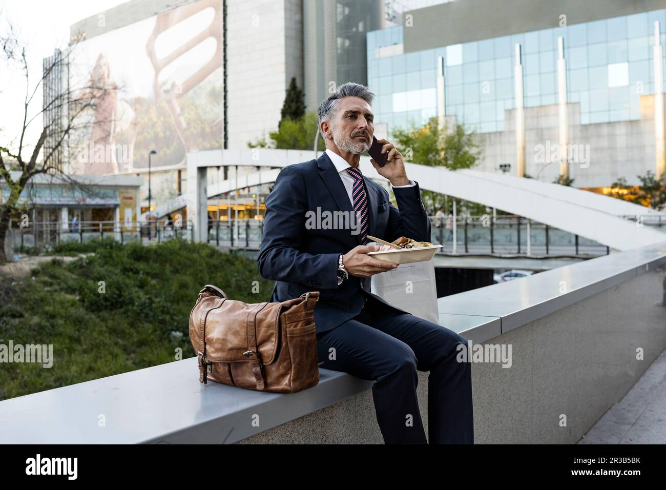 Businessman talking through smart phone holding meal sitting on wall Stock Photo