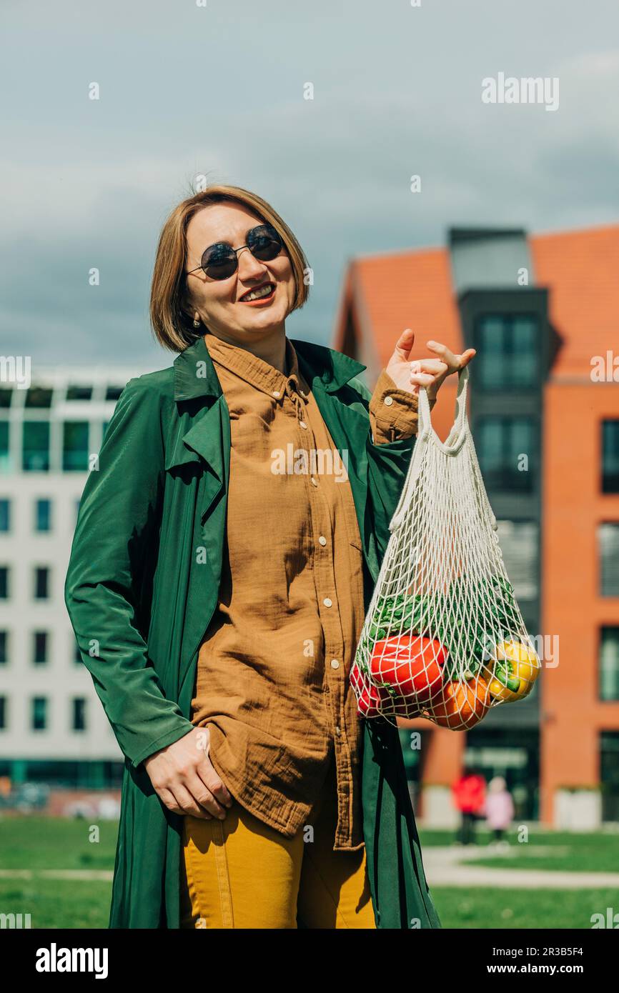Stylish woman standing with vegetables in mesh bag Stock Photo