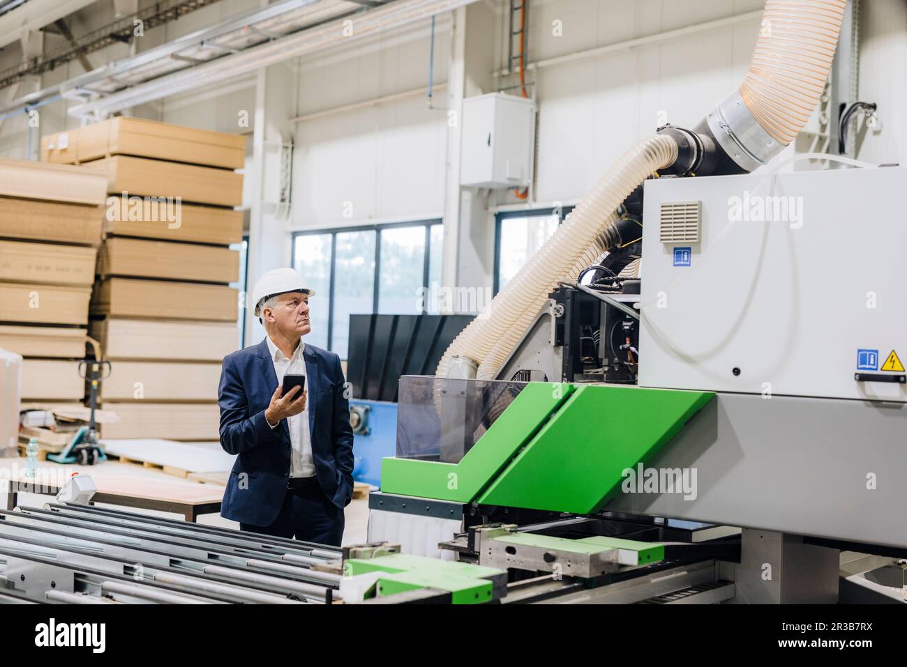 Businessman looking at modern machine holding smart phone in factory ...