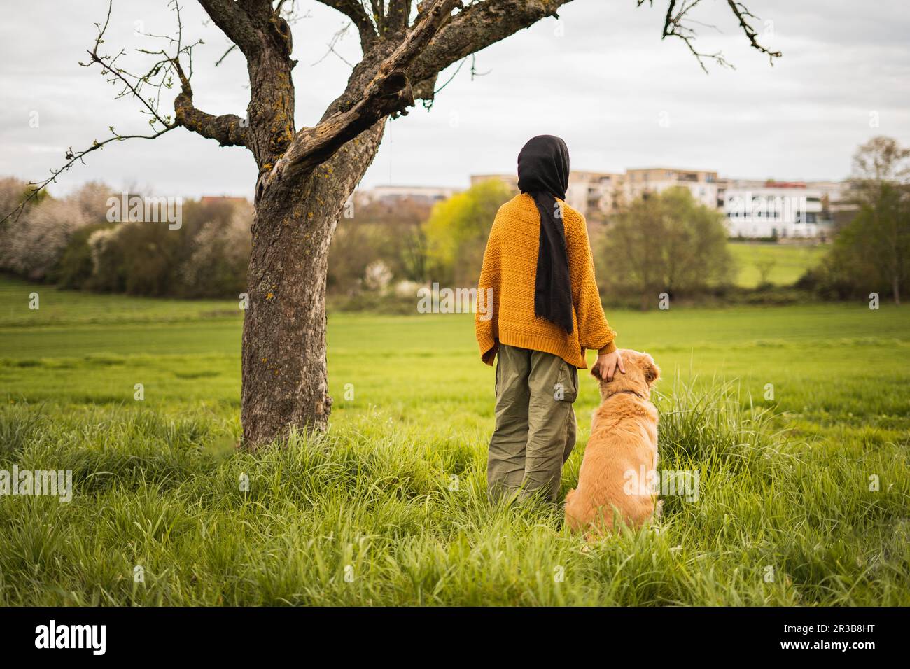 young muslim girl standing with a dog, two best friends under a tree in a park while watching the view on a walk Stock Photo