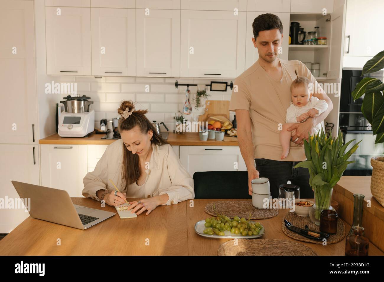 Woman working on laptop by man carrying daughter doing chores at home Stock Photo