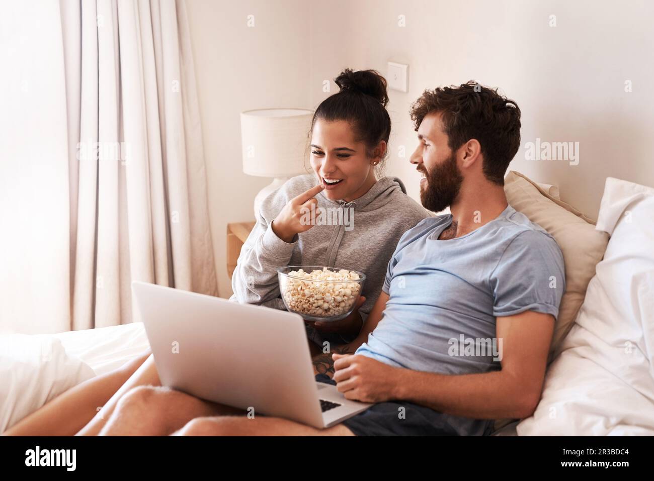 Laptop, watching movie and popcorn with couple in bed for streaming, relax or internet. Subscription, happy and love with man and woman eating at home Stock Photo