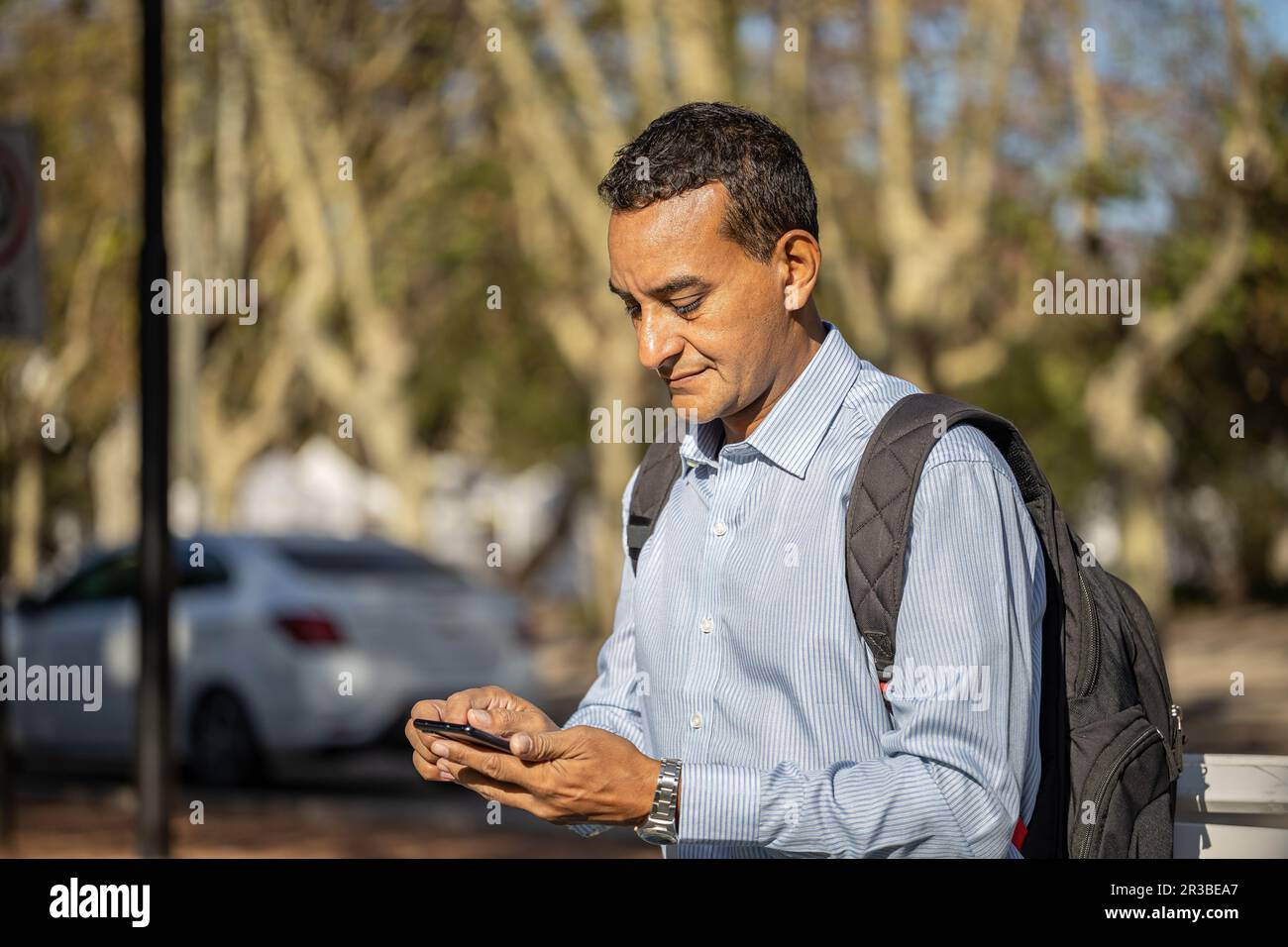 Young latin man sitting on a square bench using his mobile phone. Stock Photo