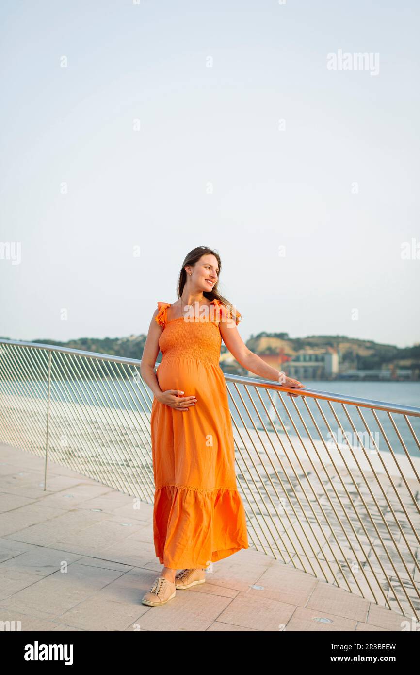 Smiling pregnant woman with hand on stomach standing by railing Stock Photo