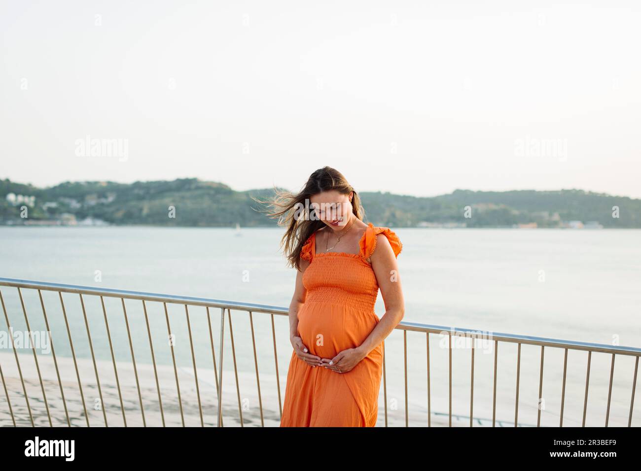 Smiling pregnant woman with hands on stomach by railing Stock Photo