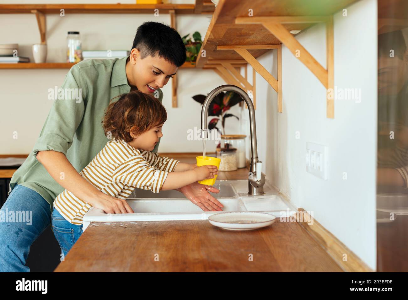 Son filling water in glass through faucet by mother in kitchen at home Stock Photo