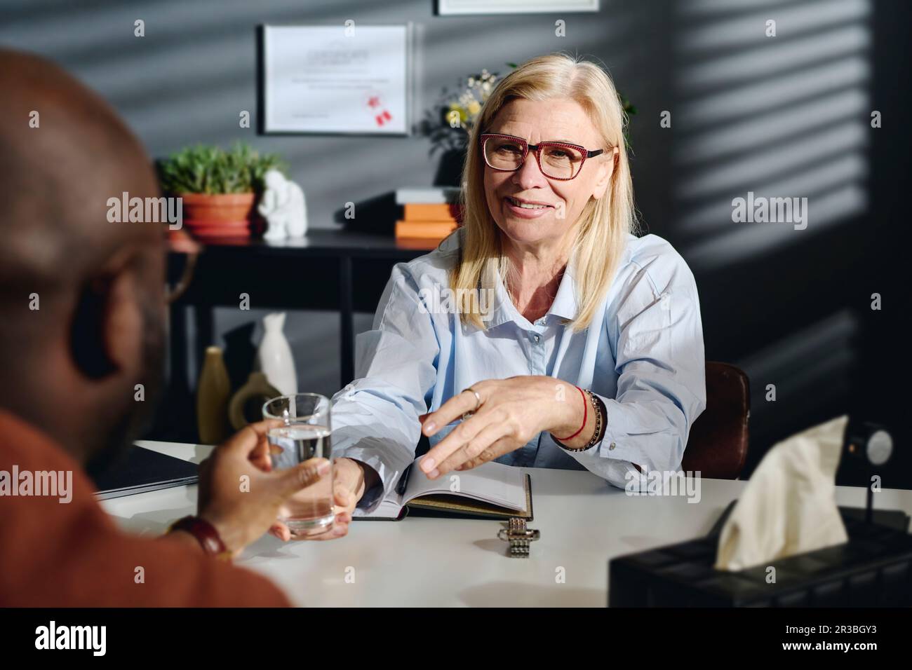 Smiling blond psychologist giving glass of water to patient at office Stock Photo