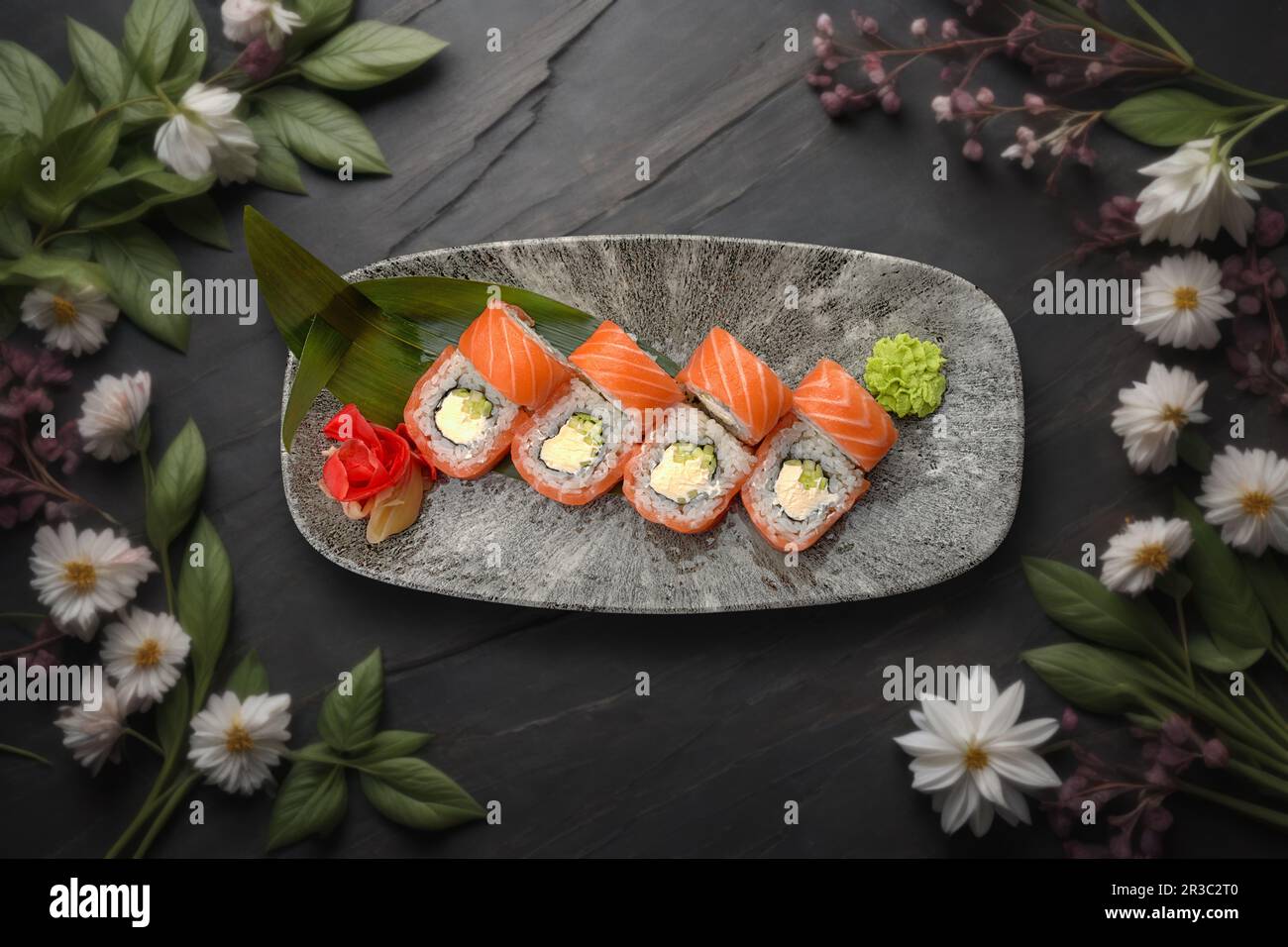 sushi roll featuring salmon, creamy cheese, and avocado on a textured stone surface with floral embellishments Stock Photo