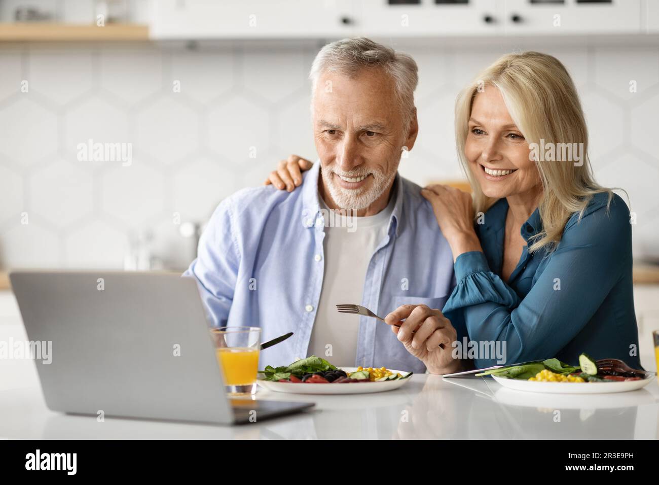 Smiling Elderly Spouses Relaxing With Laptop While Having Lunch In Kitchen Stock Photo