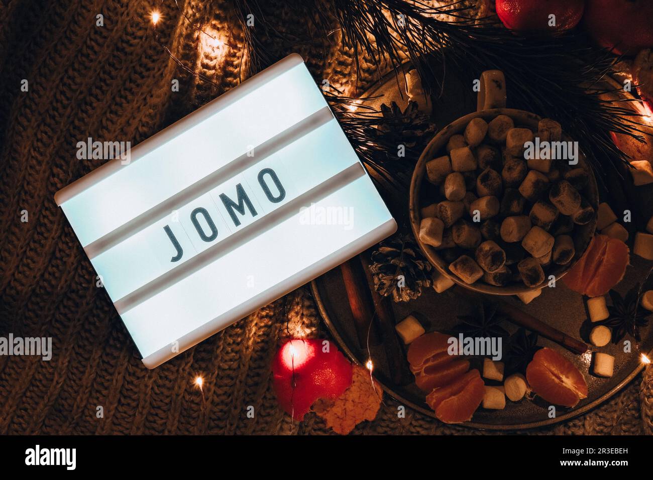 Lightbox with text JOMO Joy of missing out Cup with hot winter cacao and marshmallows tangerines spruce branch on bed. Christmas Stock Photo