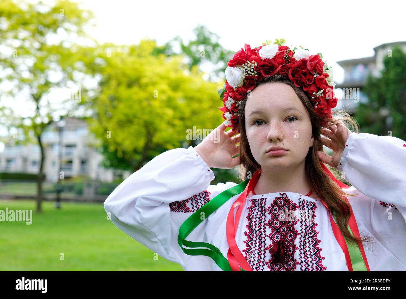 beautiful Strong brave Ukrainian young woman in red wreath of flowers of embroidered shirt with ribbons in hair stands against background of green trees looks into distance seriously power Glory Stock Photo