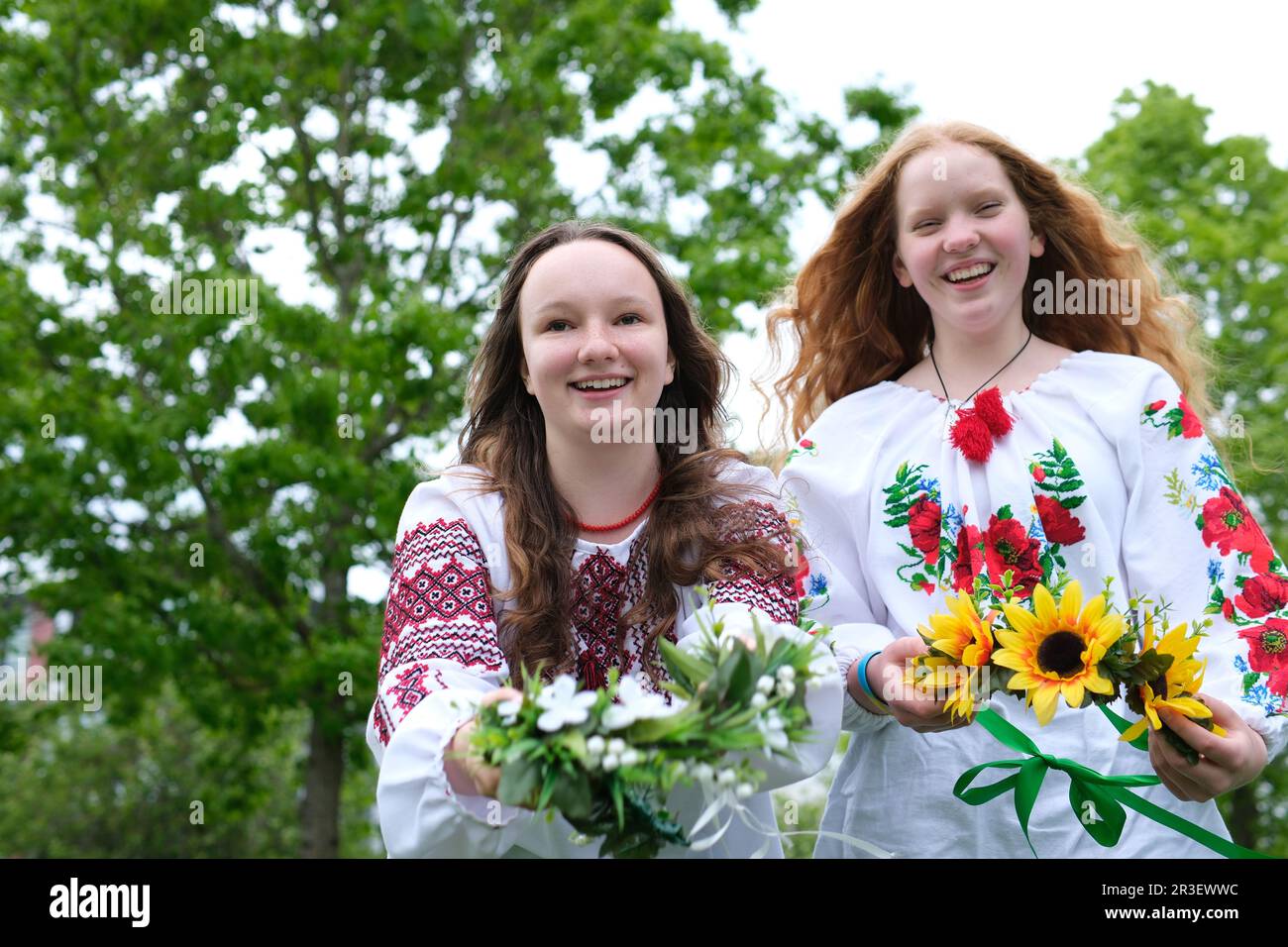 beautiful young girls wearing embroidered shirts laughing smiling putting wreaths on water facing camera stretching out wreaths to us different sunflowers Field flowers girl has red bright sunny hair Stock Photo