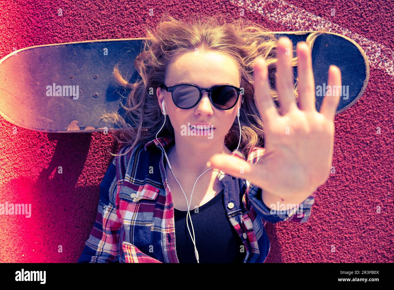 Millennial hippie Woman with sunglasses and headphones lying down on a skateboard on street. Skater girl on a longboard. Cool fe Stock Photo