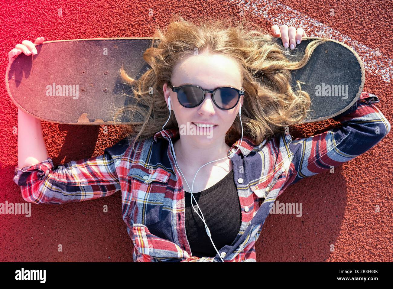 Millennial hippie Woman with sunglasses and headphones lying down on a skateboard on street. Skater girl on a longboard. Cool fe Stock Photo