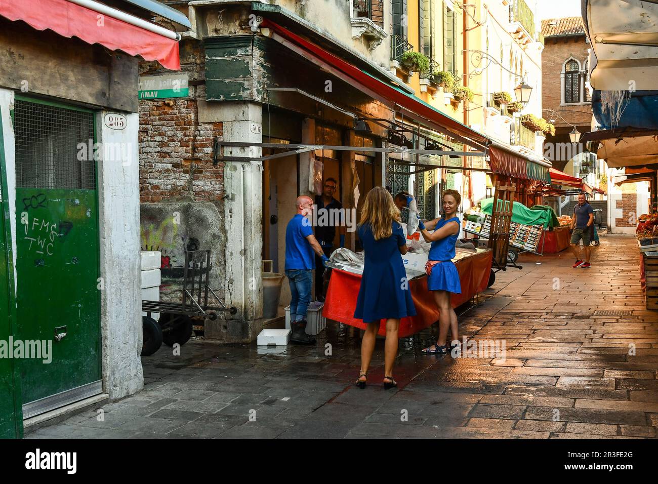 Two young foreign tourists are jokingly chatting with a fishmonger who is selling dogfish at the Fish Market of Rialto in summer, Venice, Veneto Stock Photo