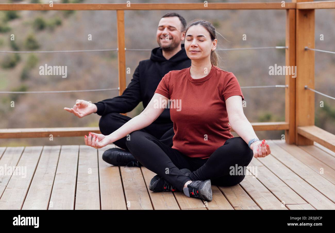 Happy relaxed man and woman doing yoga outdoors. Smiling sportsman sitting in lotus pose. Young male athlete training on wooden floor in the park. Stock Photo