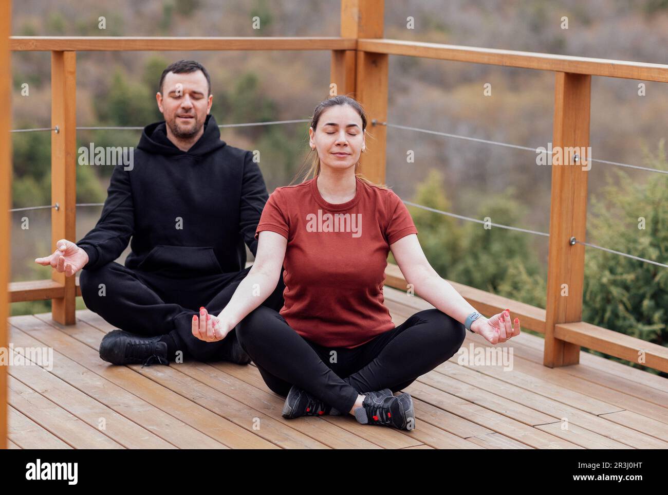Happy relaxed man and woman doing yoga outdoors. Smiling sportsman sitting in lotus pose. Young male athlete training on wooden floor in the park. Stock Photo
