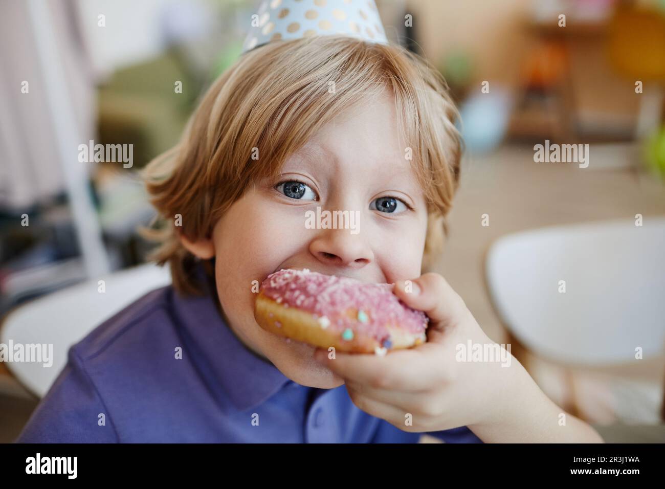 Close-up portrait of blonde little boy eating big donut during birthday party and looking at camera Stock Photo