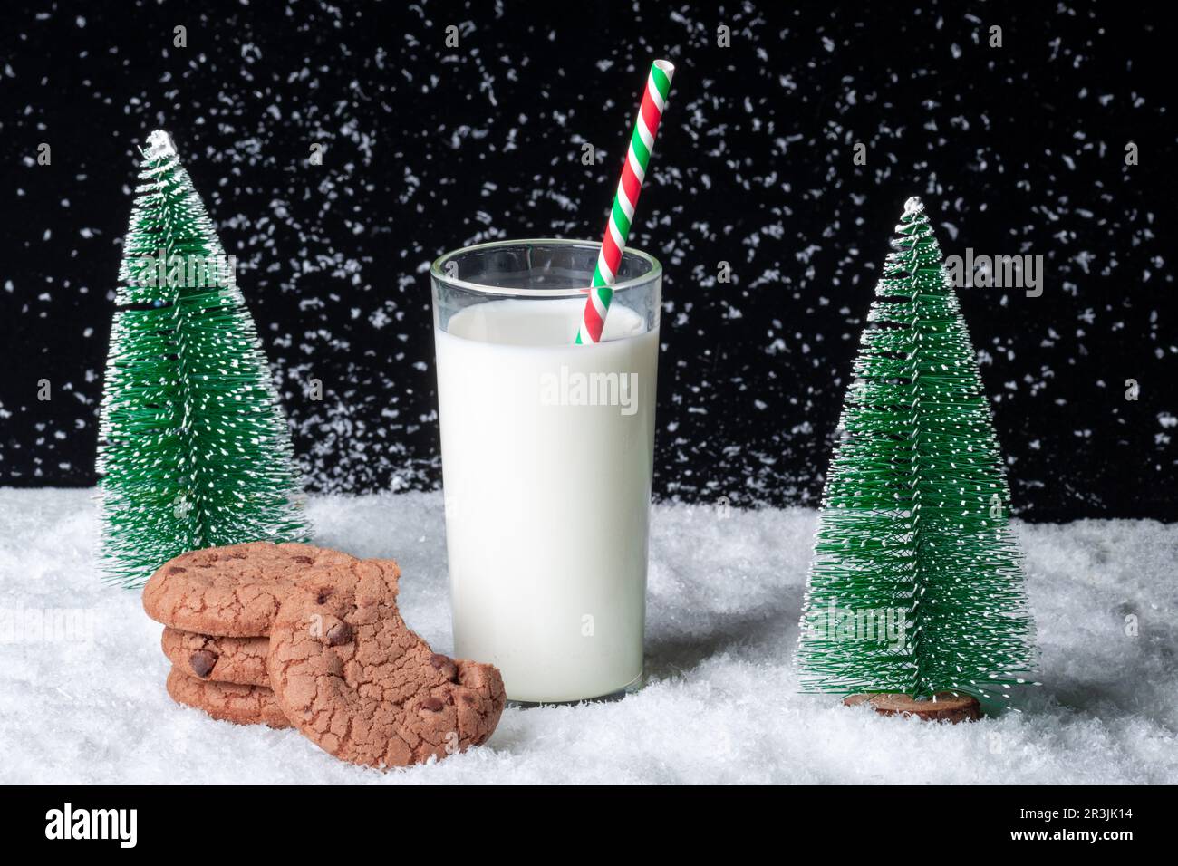 A gift for Santa. A glass of milk with a straw and a half-eaten cookie with chocolate chips on a snowy night background among toy Christmas trees. Wai Stock Photo