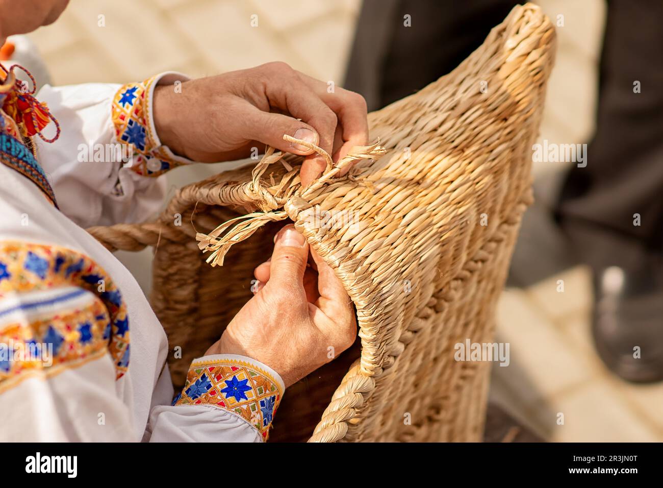 in the photo,the hands of a master in weaving products from a vine, he is weaving a laundry basket, he is wearing a shirt sewn and embroidered by hand Stock Photo