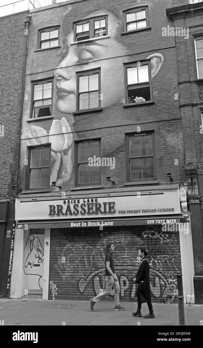 Pedestrians walk past a large face mural on a building facade along Brick Lane East London,captured in black and white and highlighting everyday life Stock Photo