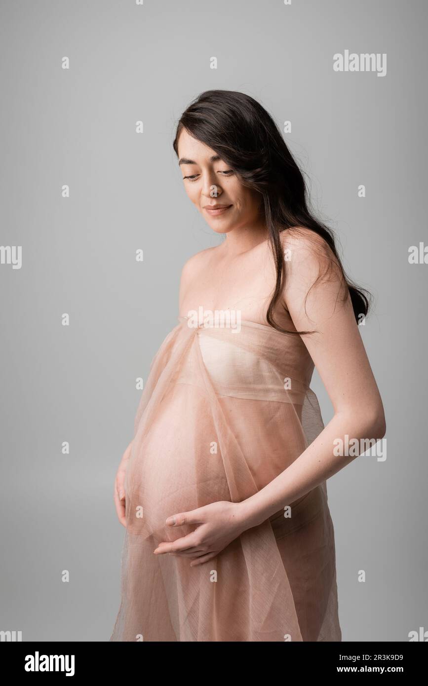 charming brunette mother-to-be in airy chiffon cloth and golden bracelets touching belly and smiling isolated on grey background, maternity fashion co Stock Photo